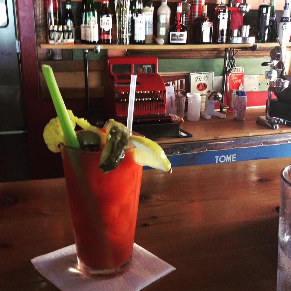 A Bloody Mary garnished with celery, a pickle and a lemon wedge on the wood bar, a vintage red cash register behind it.