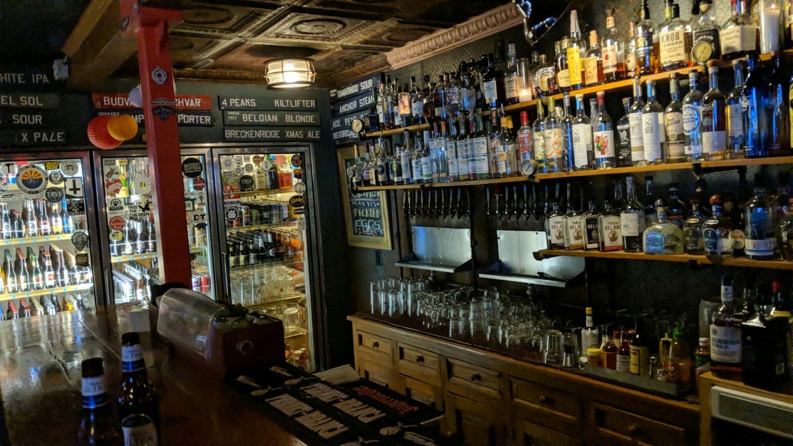 The back bar under a pressed-tin ceiling, shelves packed with bottles and a row of beer-name signs above the taps.