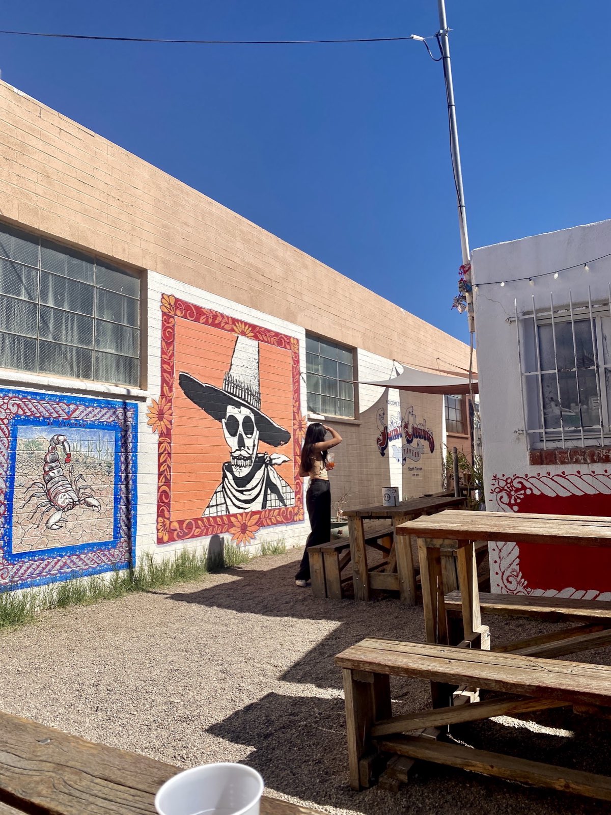 The gravel back patio in daylight with wooden picnic benches and painted murals of a scorpion and a skeleton in a wide hat.